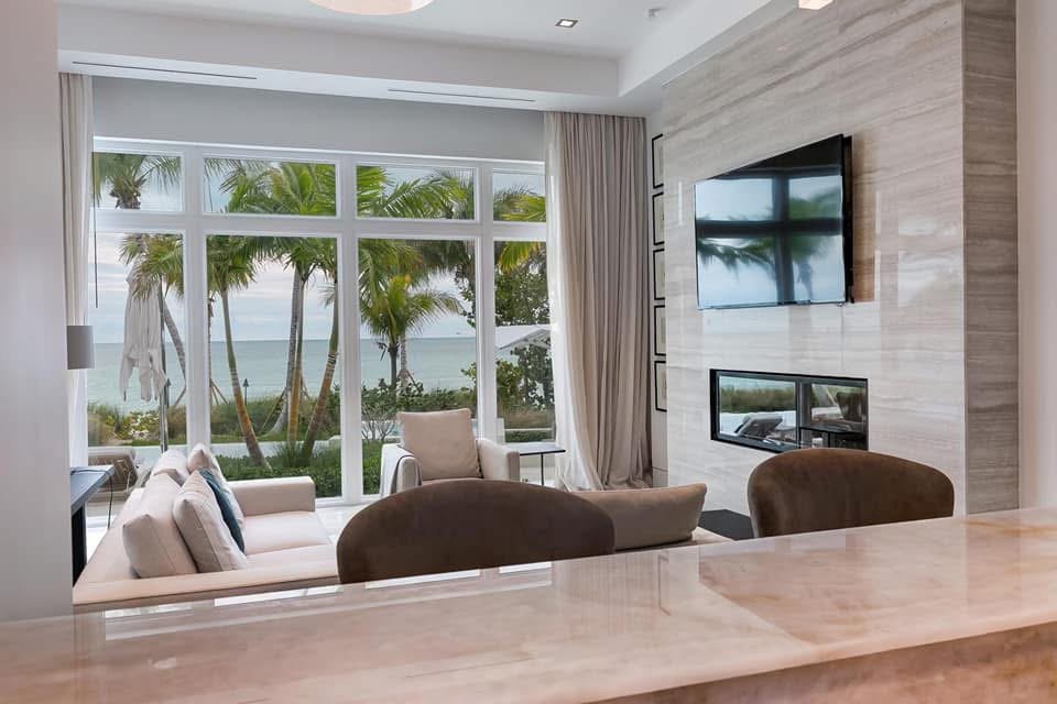 Living room at Albany framed by full height windows onto the beach, with a stone clad fireplace wall.